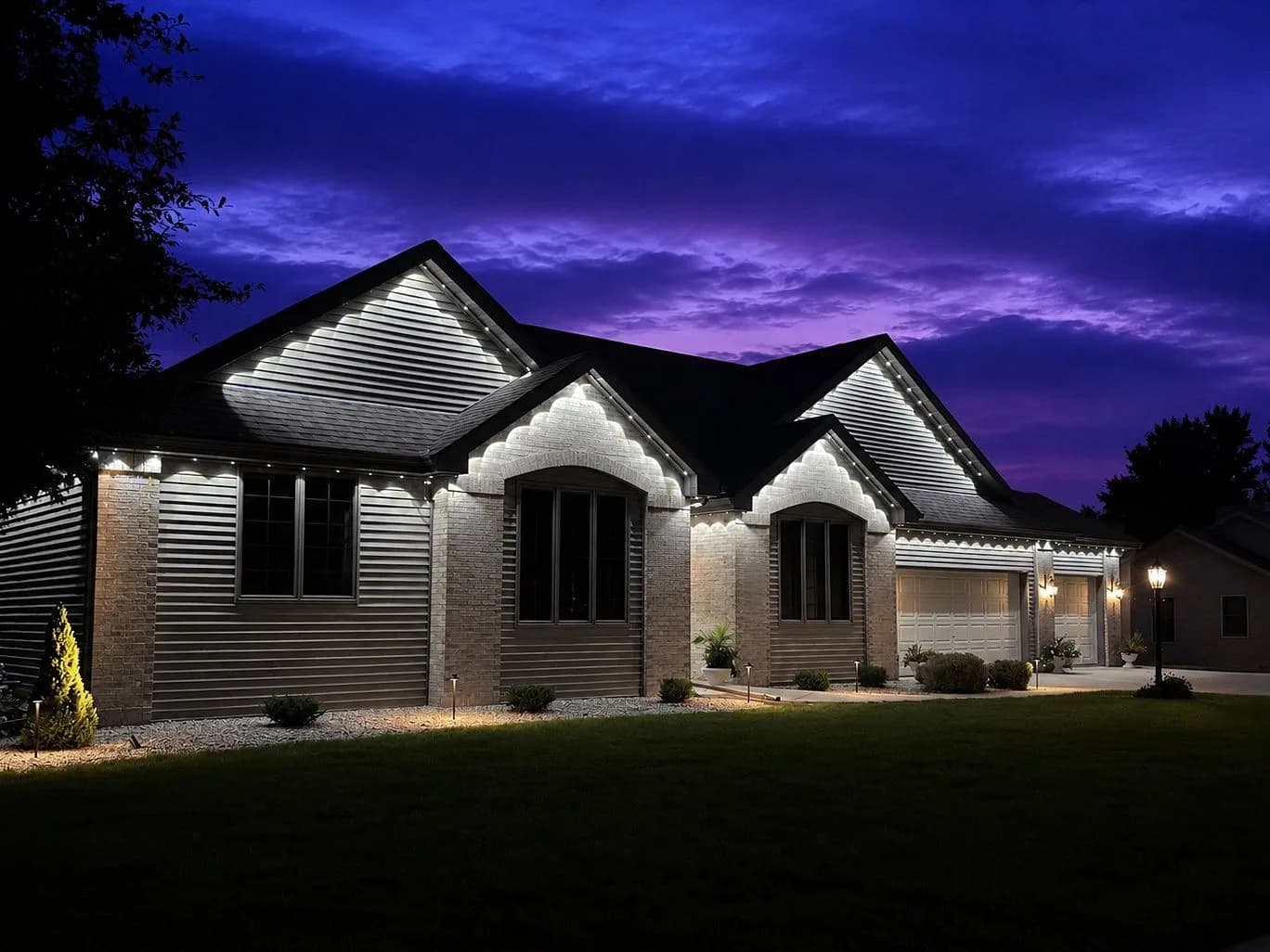 Real Kussmaul Co. permanent lighting in cool white picking out every gable of a brick ranch under a purple dusk sky