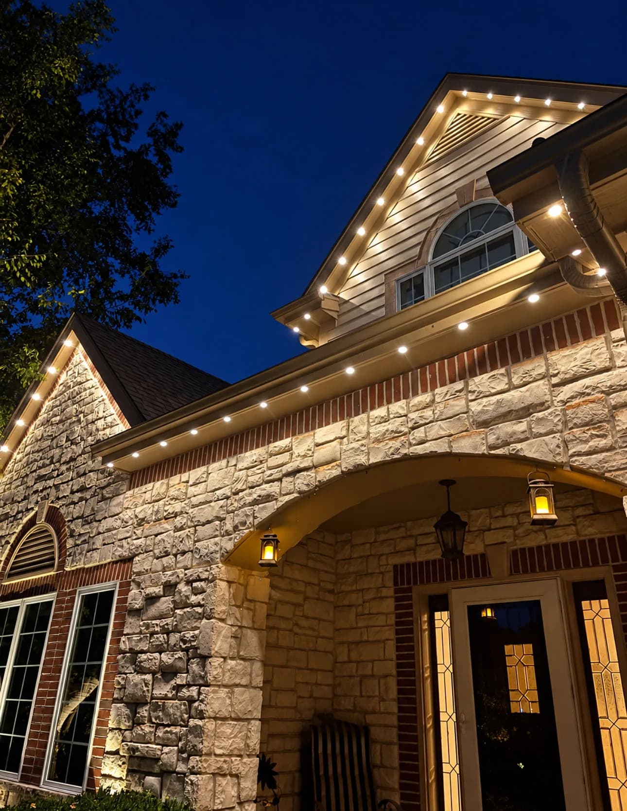Real Kussmaul Co. permanent lighting in warm white along the gable and eave above a stone-and-brick front entry at night