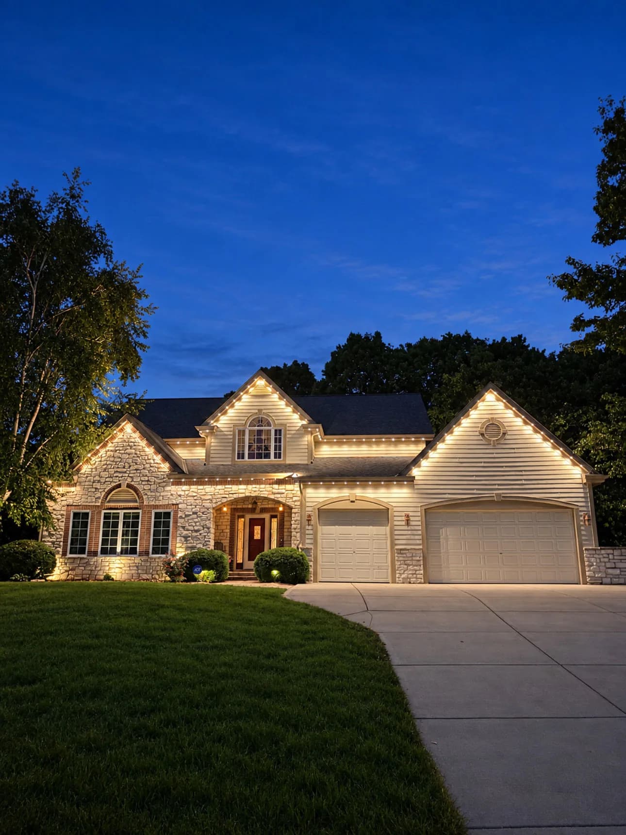 Real Kussmaul Co. permanent lighting in warm white across a stone two-story home at dusk