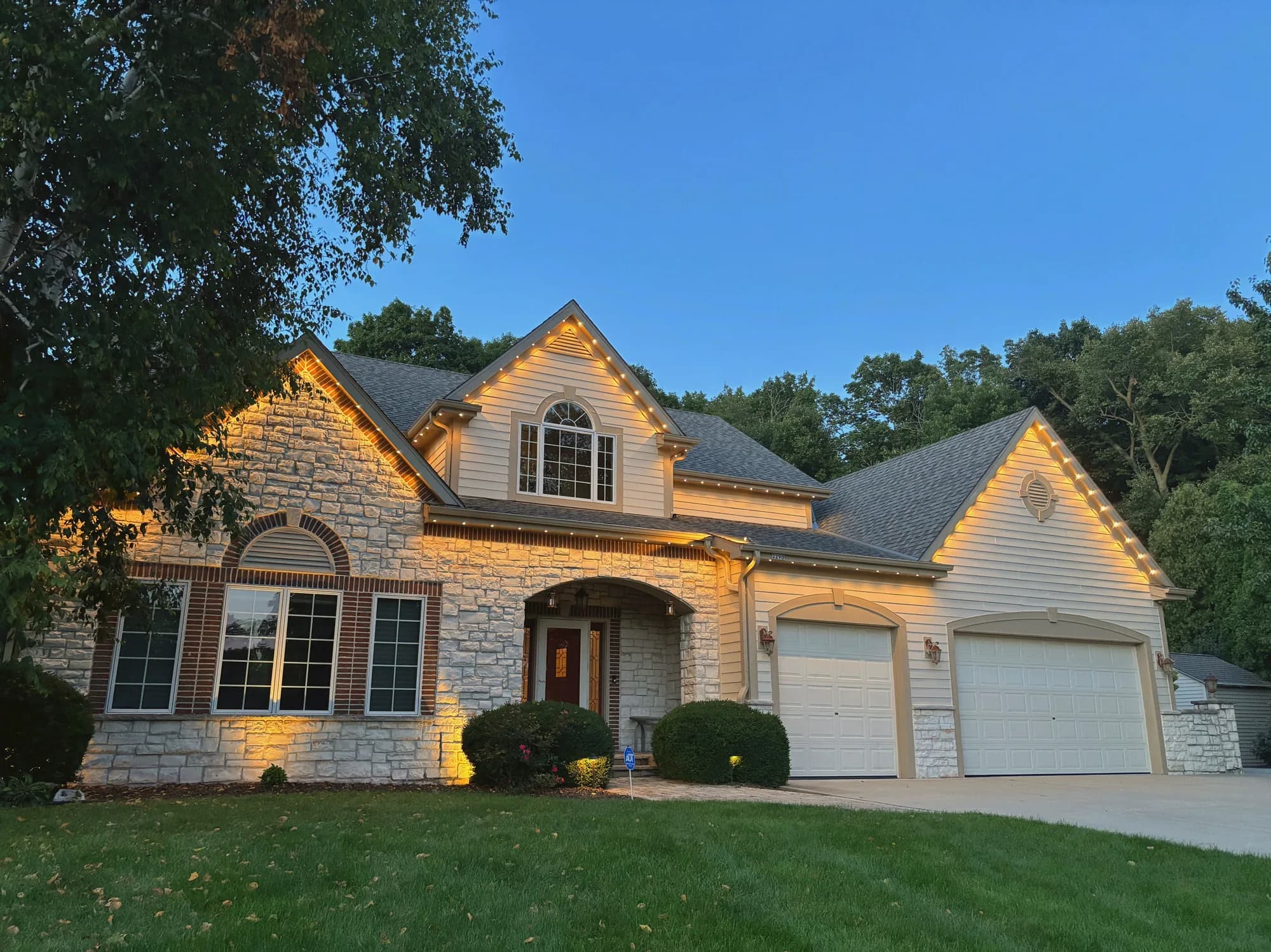 Real Kussmaul Co. permanent lighting in warm white along the gables and roofline of a two-story stone-and-siding home at golden hour