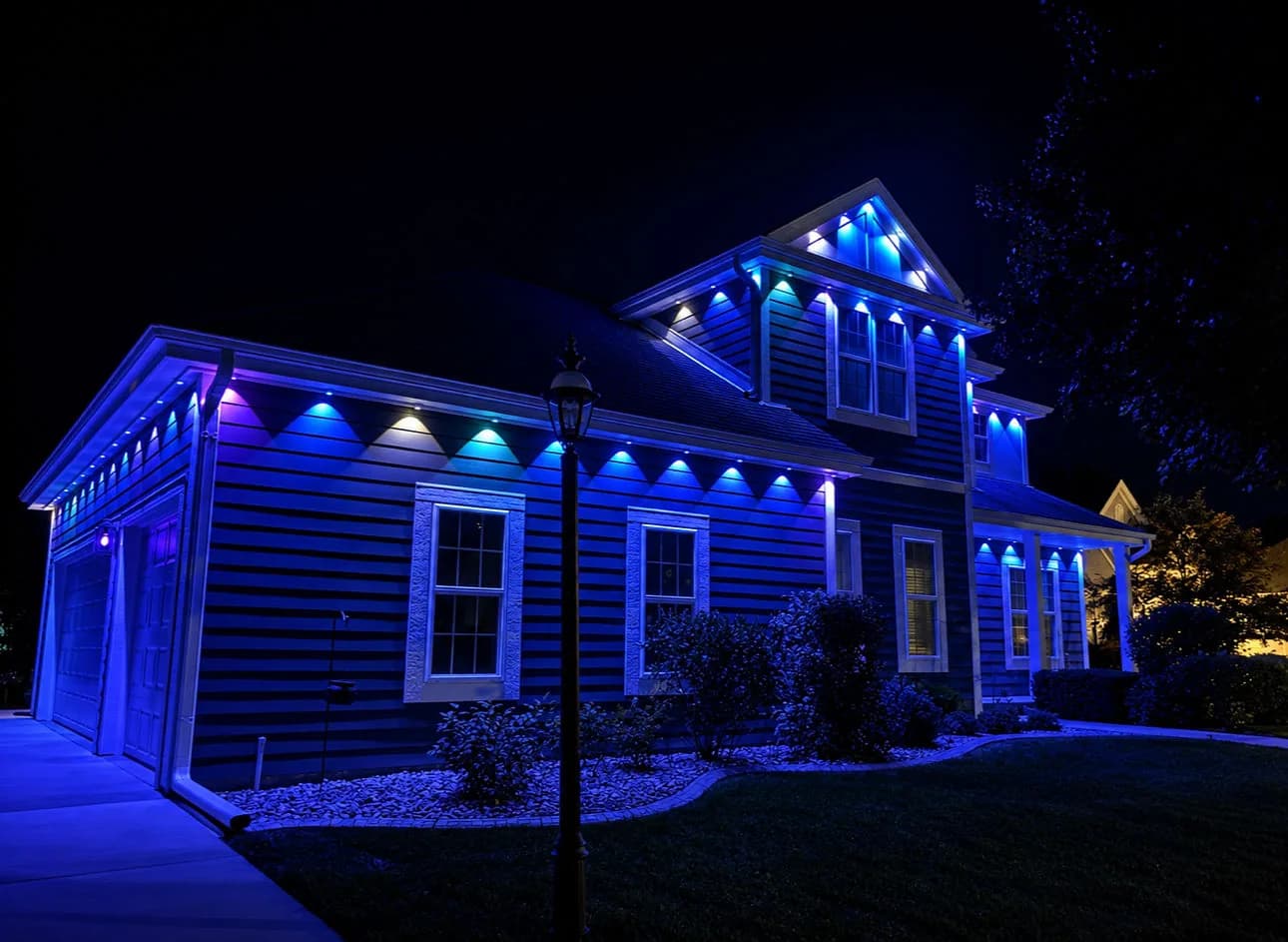 Real Kussmaul Co. permanent lighting in blue with soft purple and white accents along a two-story home's garage side