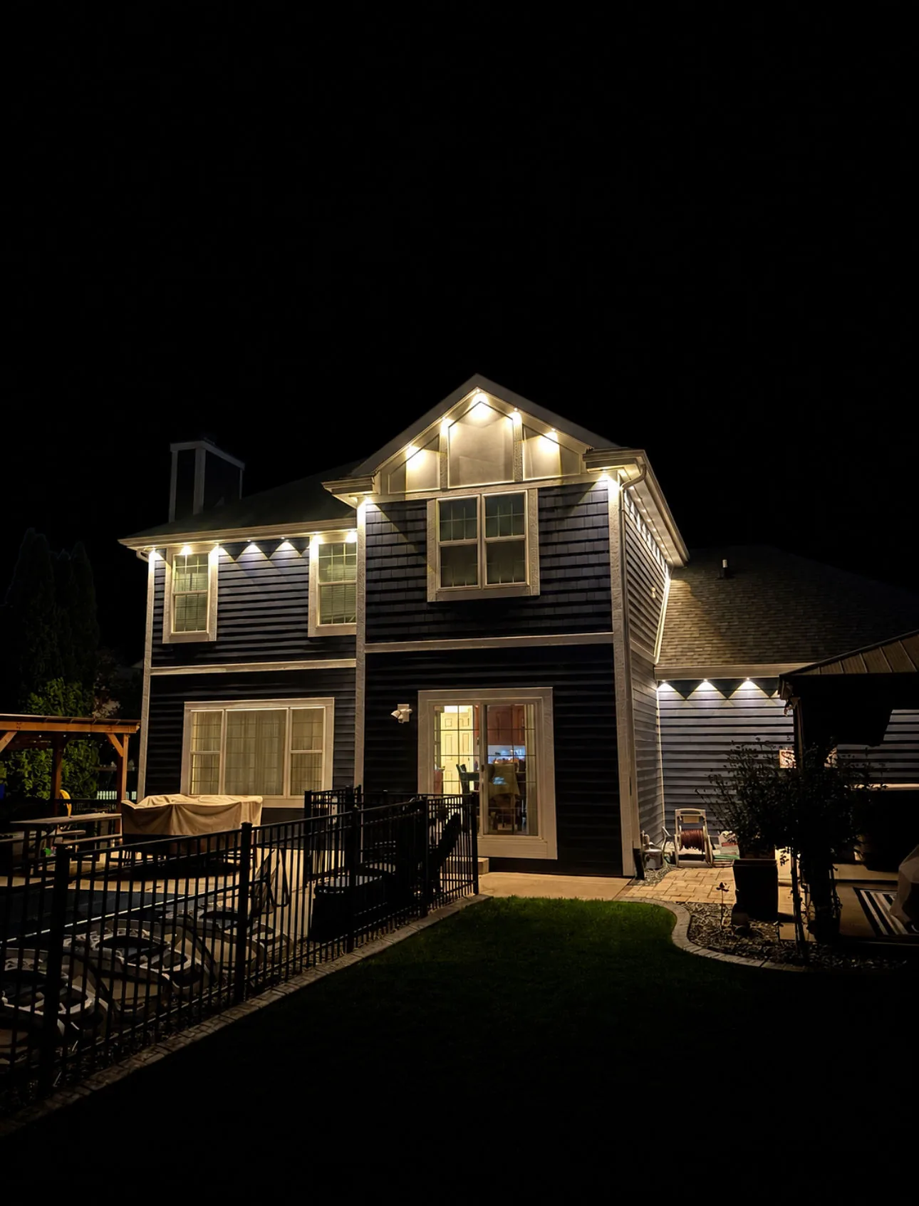 Real Kussmaul Co. permanent lighting in warm white on the back of a two-story home above a fenced pool patio