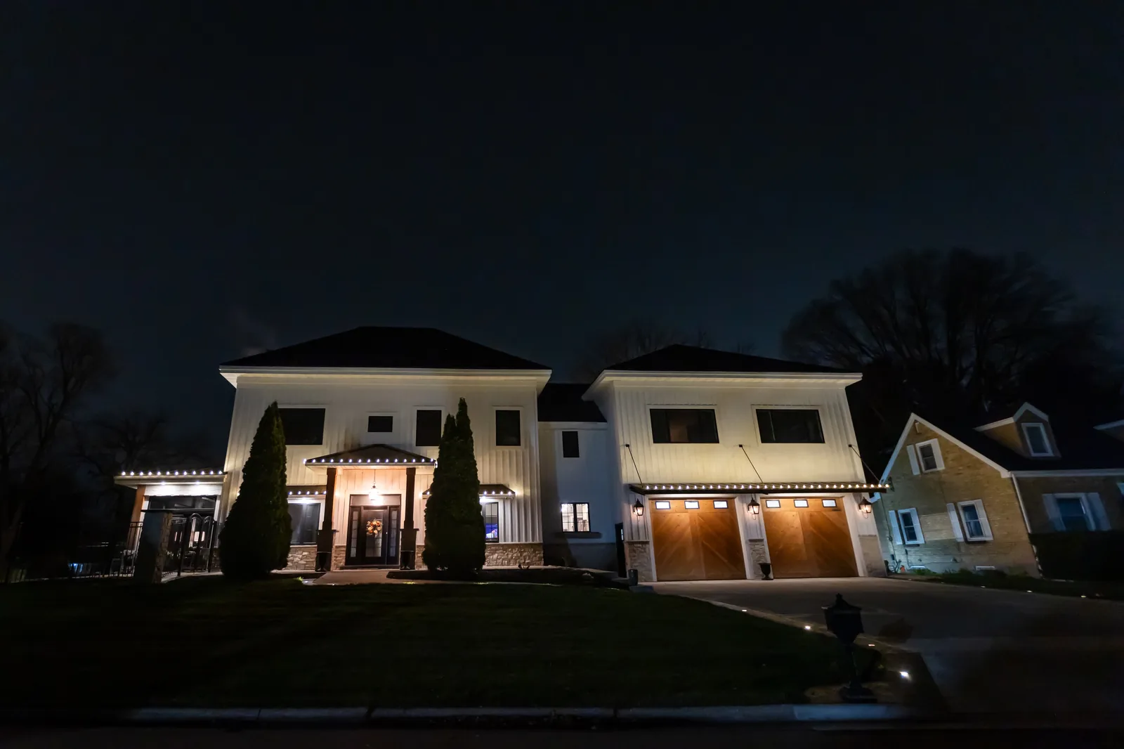 Two-story white board and batten home with warm white roofline lighting and driveway lit at dusk