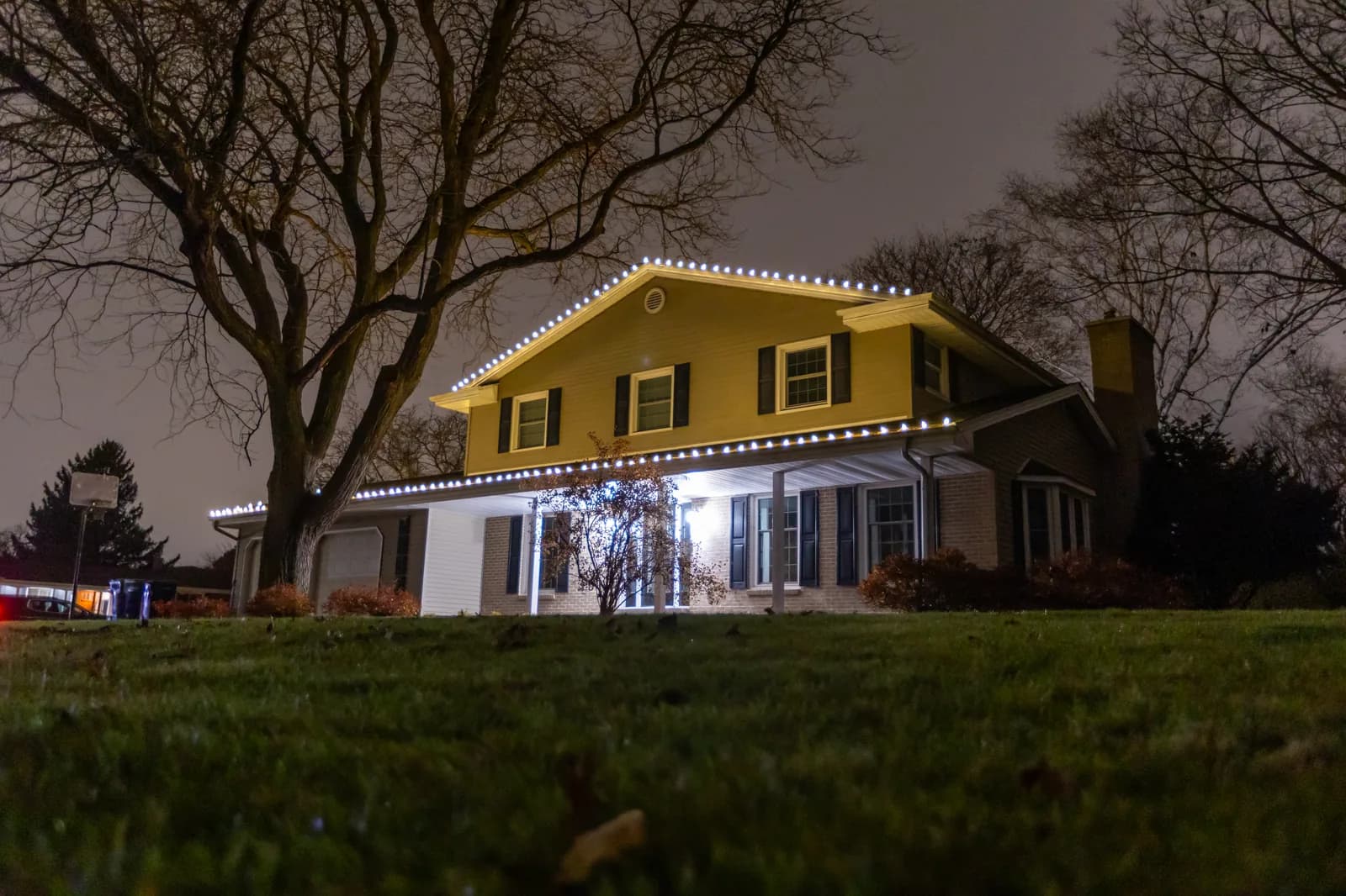 Low angle view of yellow two-story home with cool white gable and eave lighting under bare trees