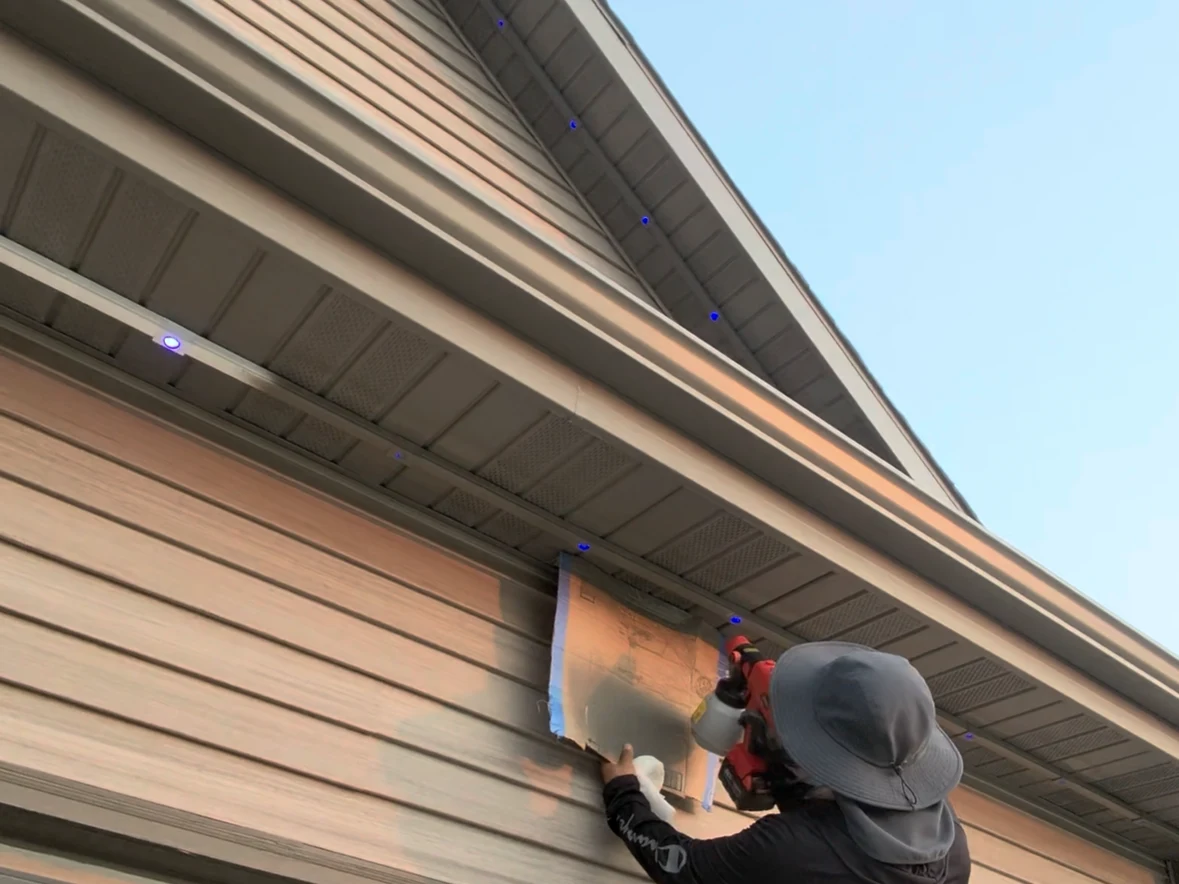 A Kussmaul Co. crew member spray-painting a masked section of lighting track to match tan siding, with the finished run above him already blended into the soffit and only its lit points visible