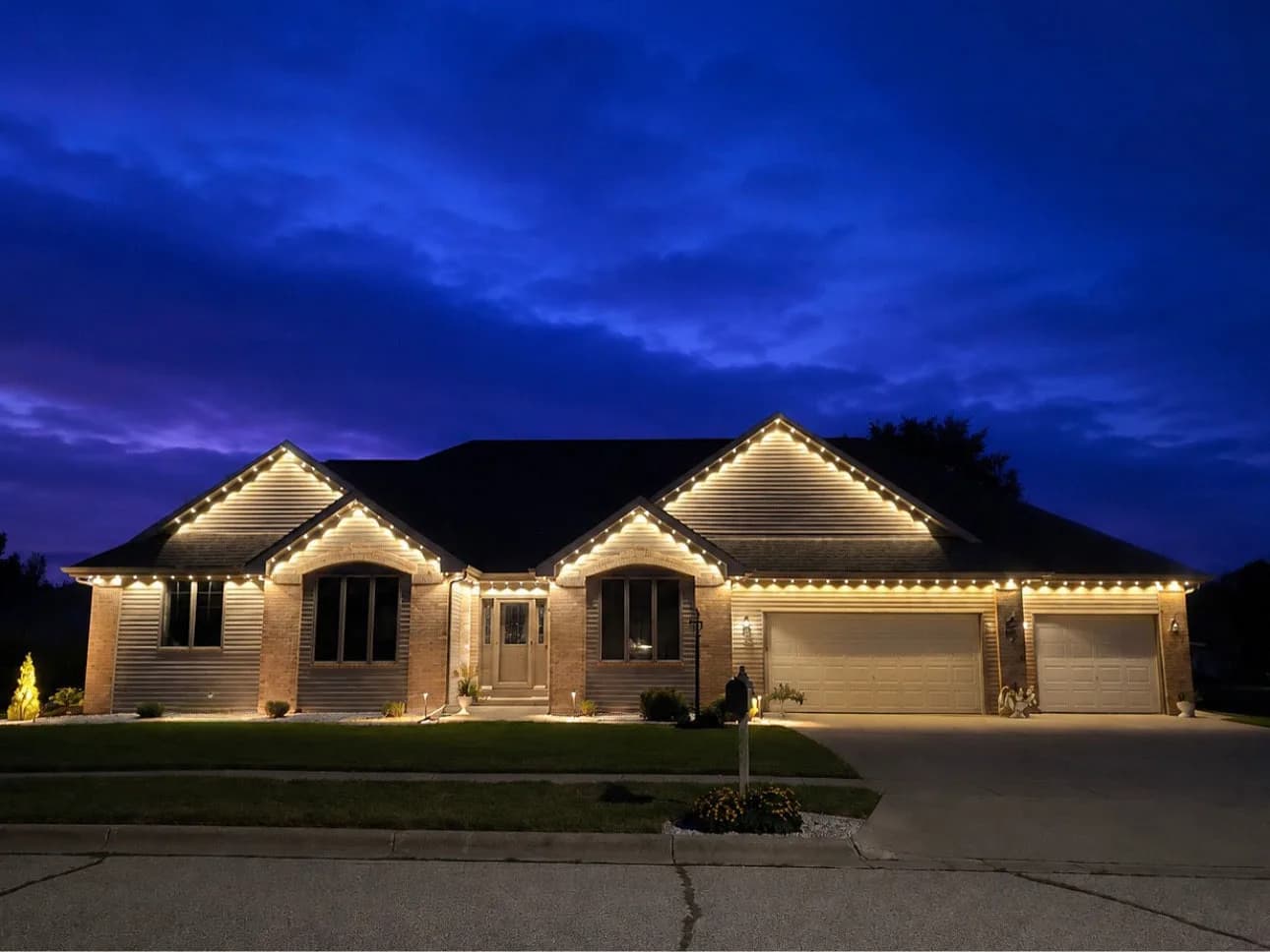The same brick ranch home with its roofline lighting set to warm white against a deep blue dusk sky.