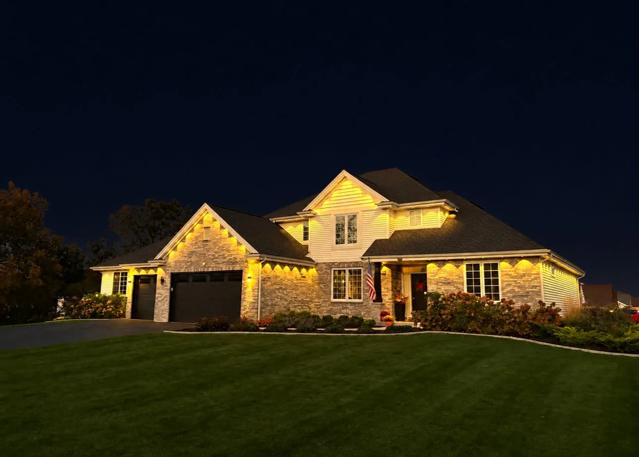 Real Kussmaul Co. permanent lighting in warm white across a brick two-story home and its front lawn, seen from the side