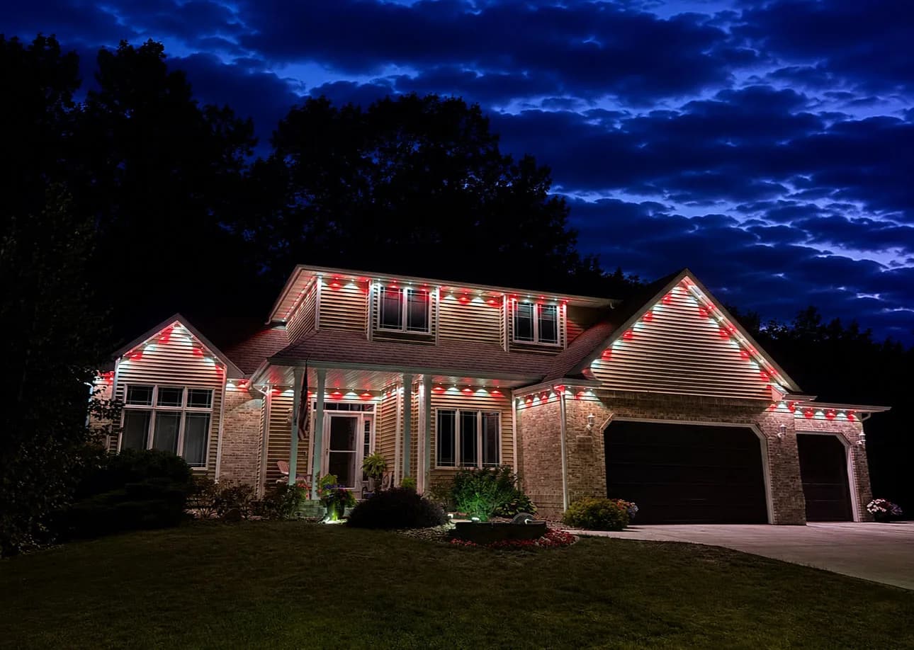Two-story home with red and white permanent lighting scene along roofline and gables at dusk