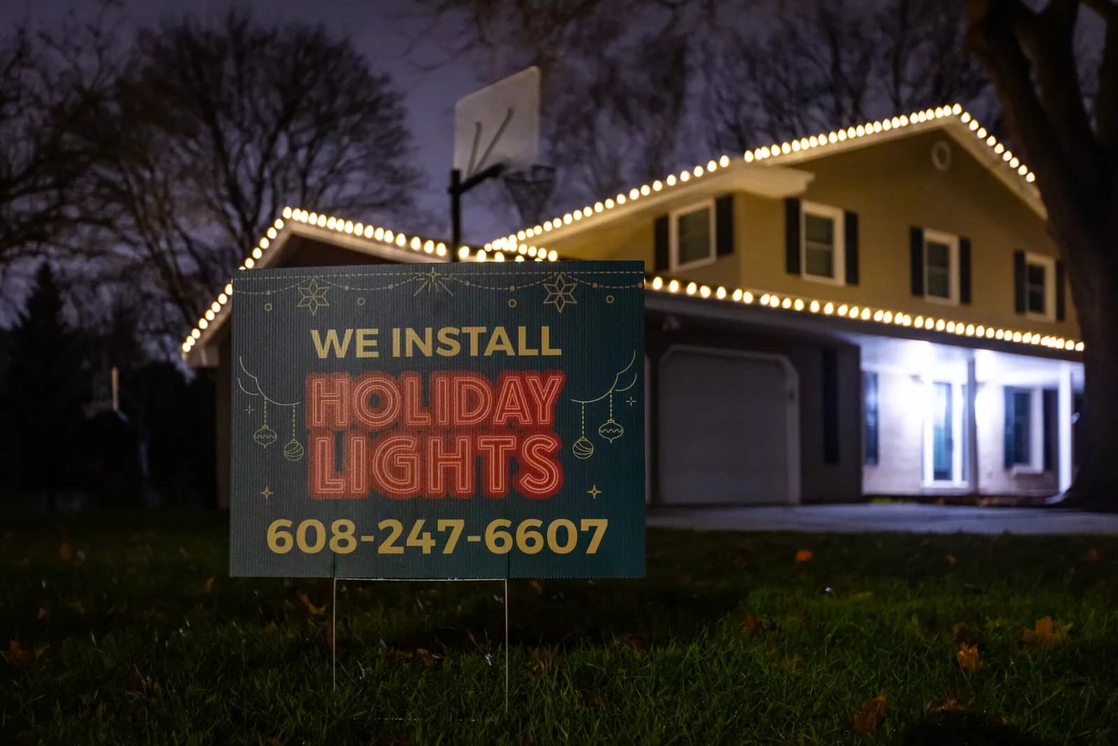A Kussmaul Co. holiday lighting yard sign in the foreground with a warmly lit two-story home glowing behind it at night
